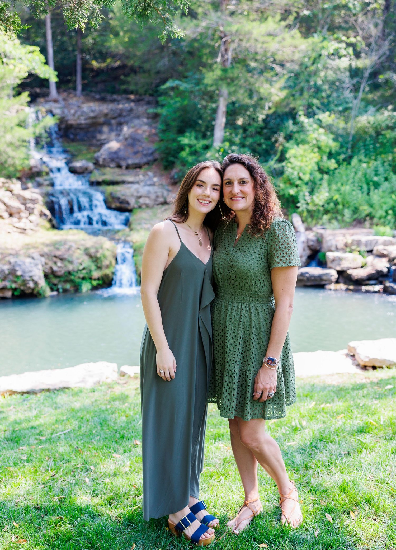 Two people stand together in green outfits by a scenic waterfall and pond surrounded by lush forest landscape.