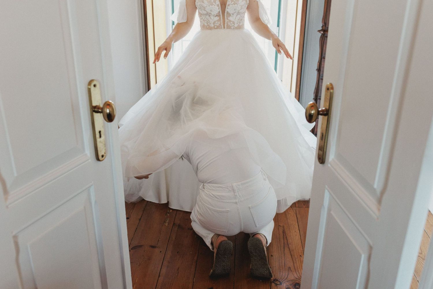 A flowing white wedding dress with tulle skirt billows over brown hardwood flooring between elegant white double doors.