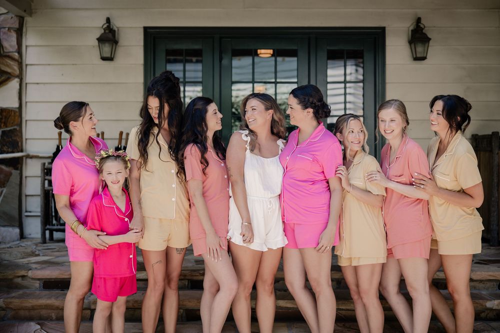 A group of bridesmaids in pink and beige dresses laugh together on a porch during a wedding celebration.