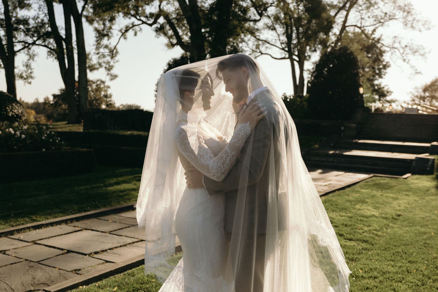 A romantic wedding couple embraces outdoors in a garden setting with flowing veils and golden sunlight.