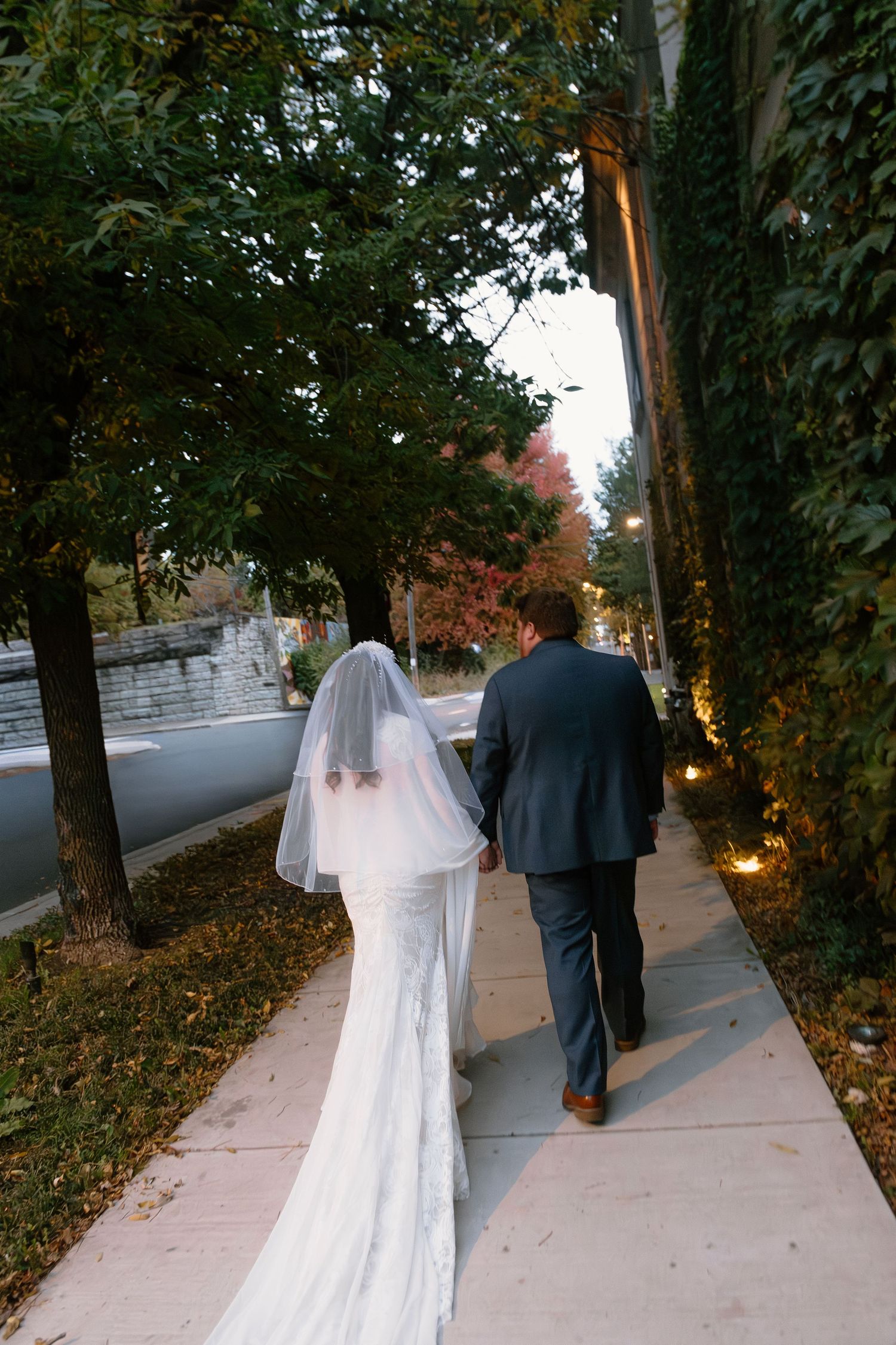 A couple in wedding attire walks down a tree-lined sidewalk during an autumn evening.