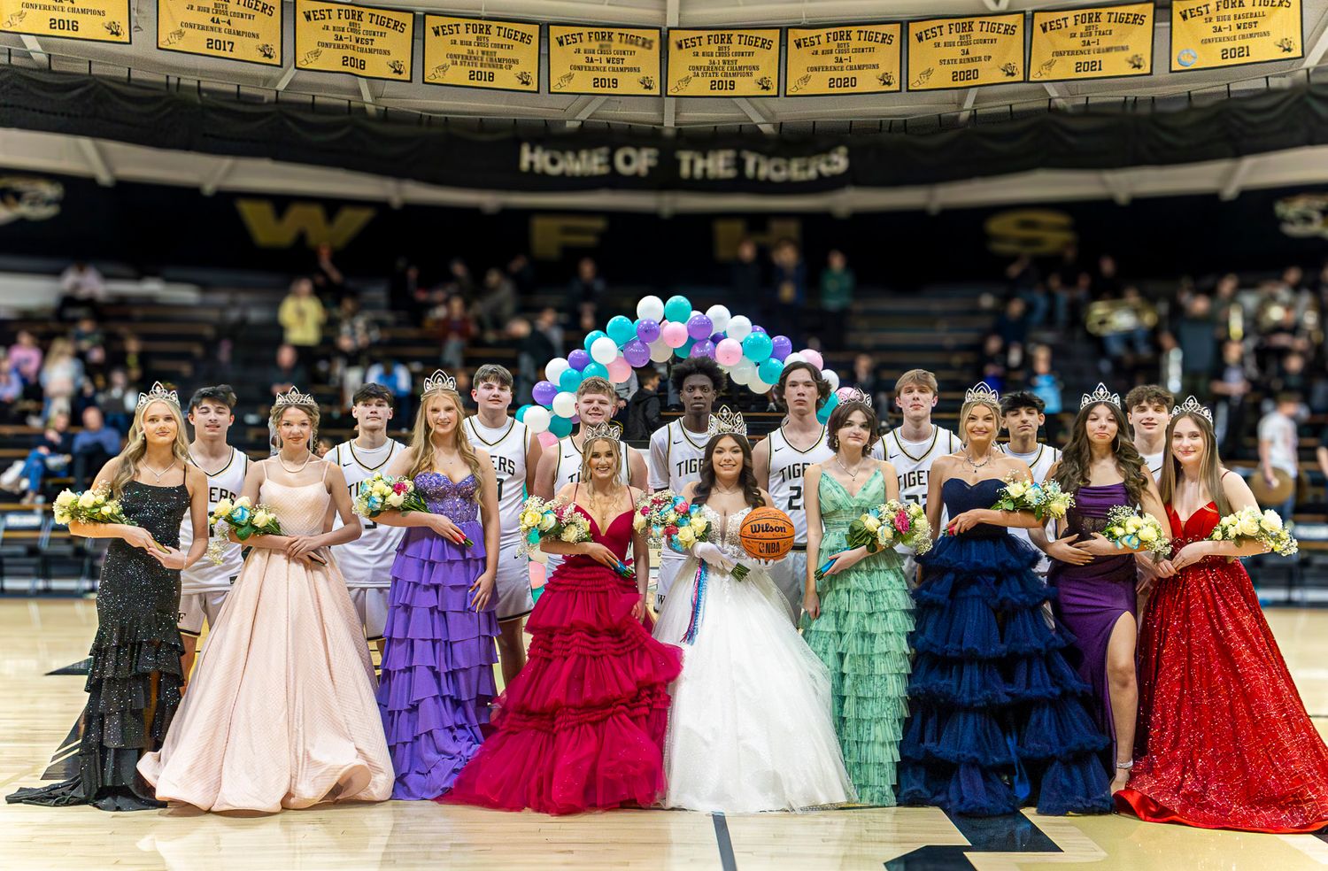 A line of women in colorful formal gowns stand under a balloon arch at an indoor event venue.