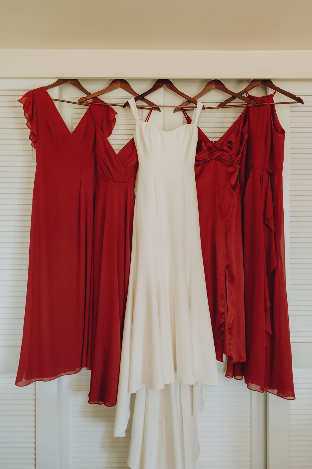 Wedding party attire hanging on wooden hangers featuring red bridesmaid dresses and a white wedding gown.