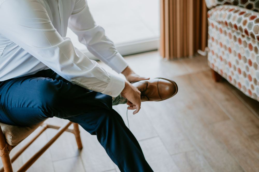 Wedding preparation scene showing dress shoes and formal attire being put on in a sunlit room.