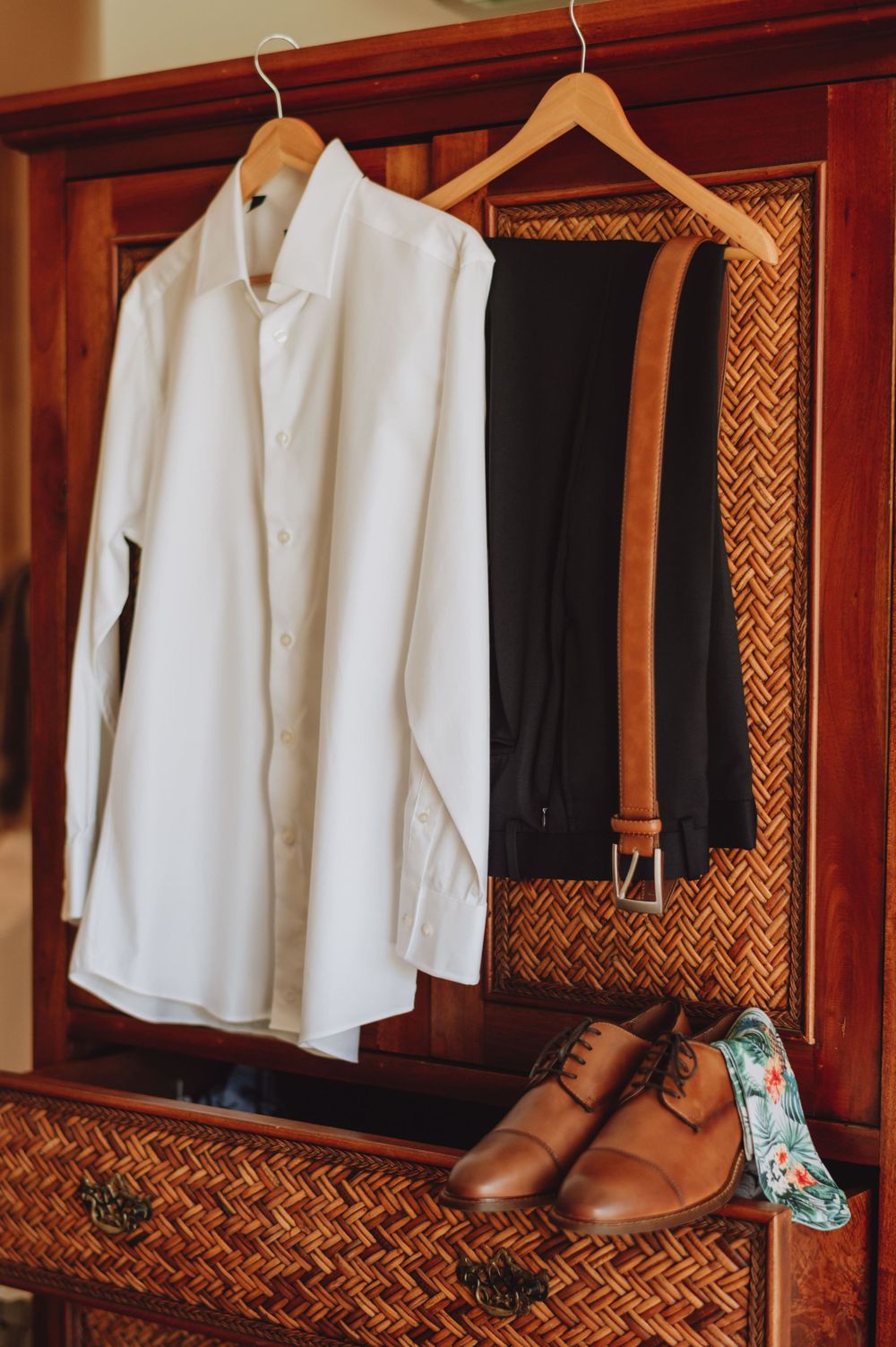 Men's formal attire and brown leather shoes displayed in wooden wardrobe with woven panels.