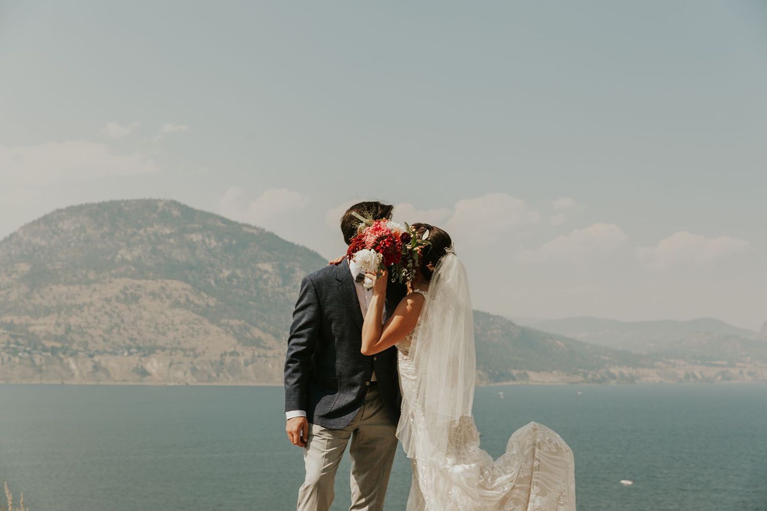 Bride and groom kissing with flowing veil and bridal bouquet against a breathtaking lake and mountain view.