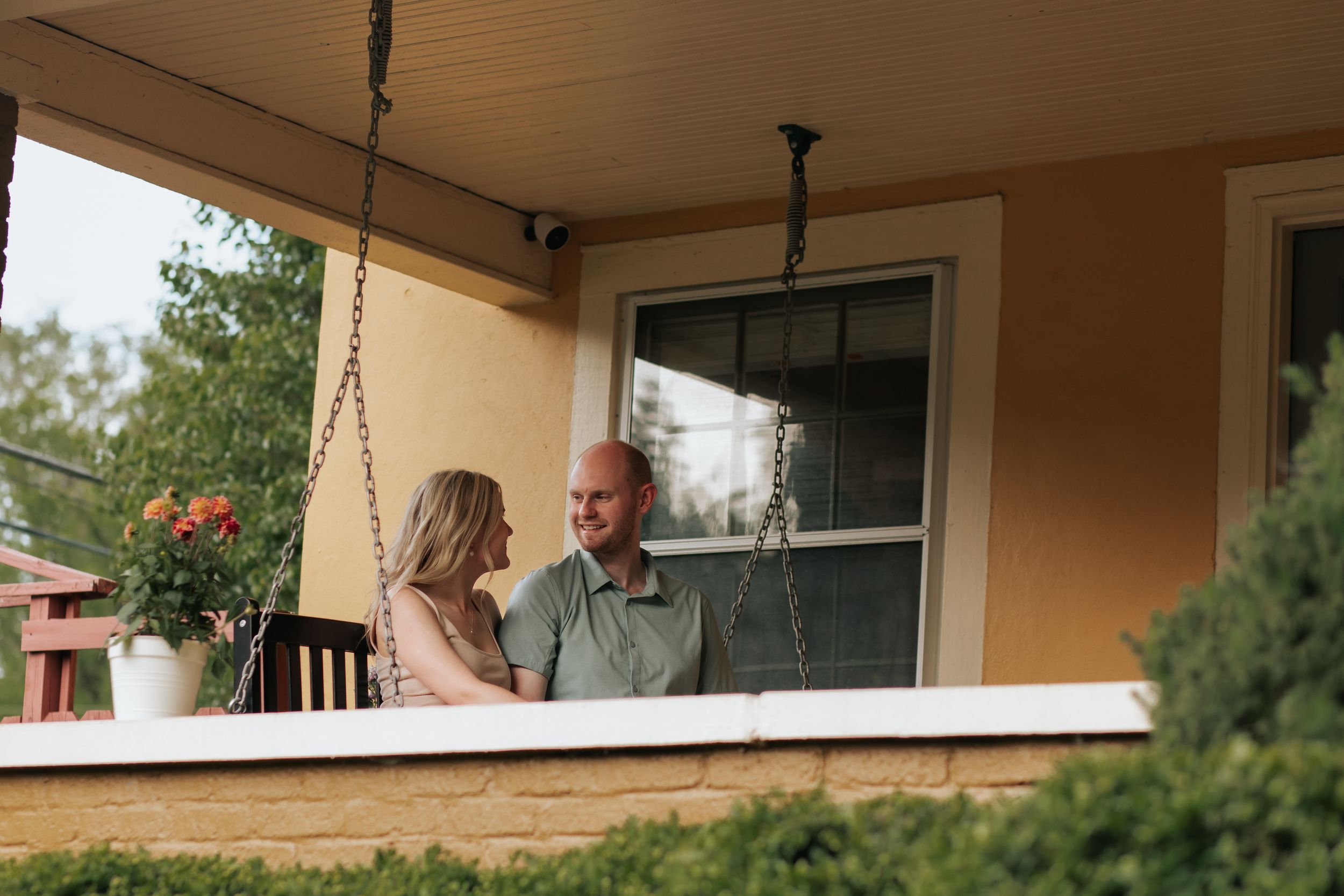 Two people sit and chat on a covered front porch with beige walls and white trim on a sunny day.