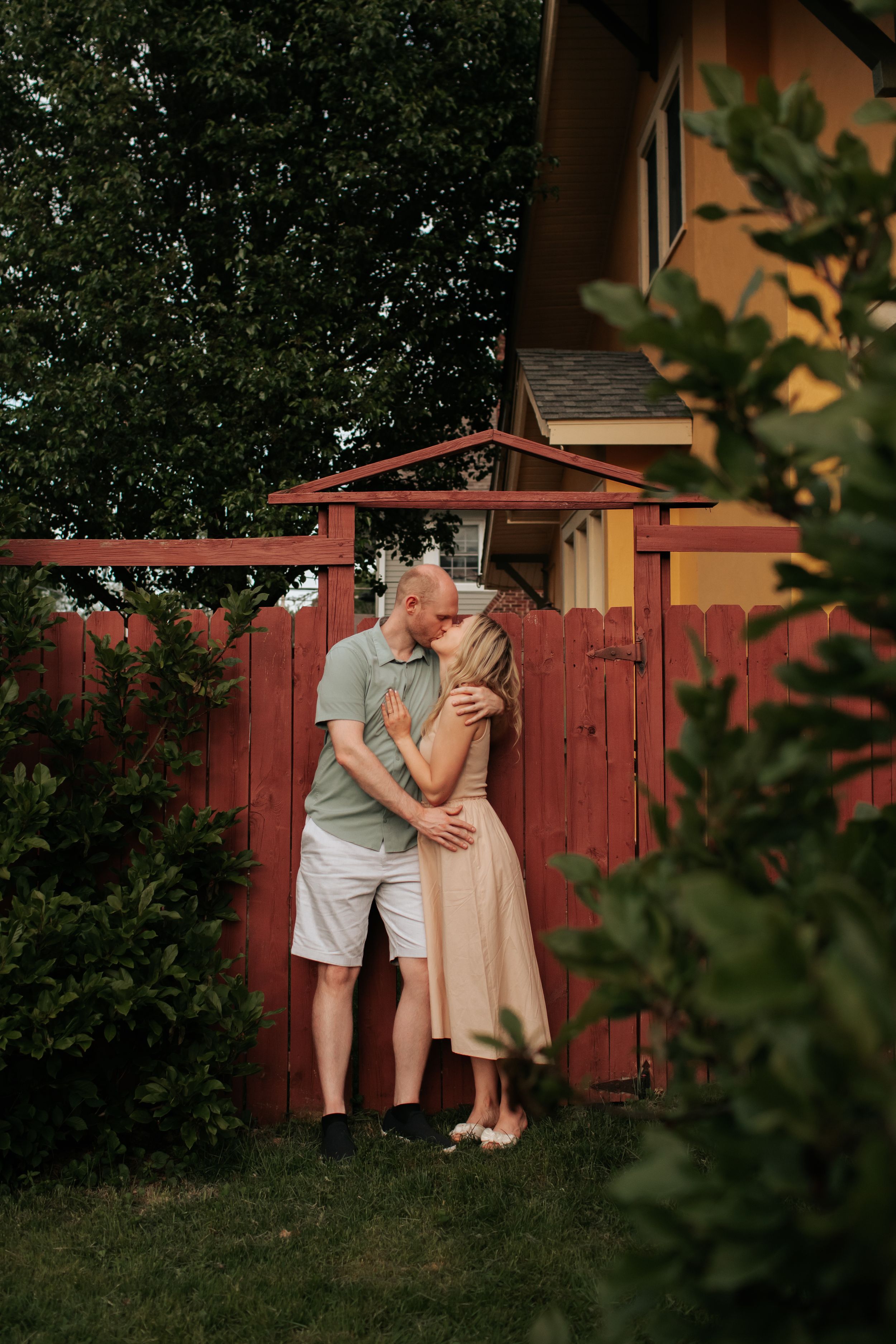 A couple shares an intimate moment together against a rustic red wooden fence in a garden setting at sunset.