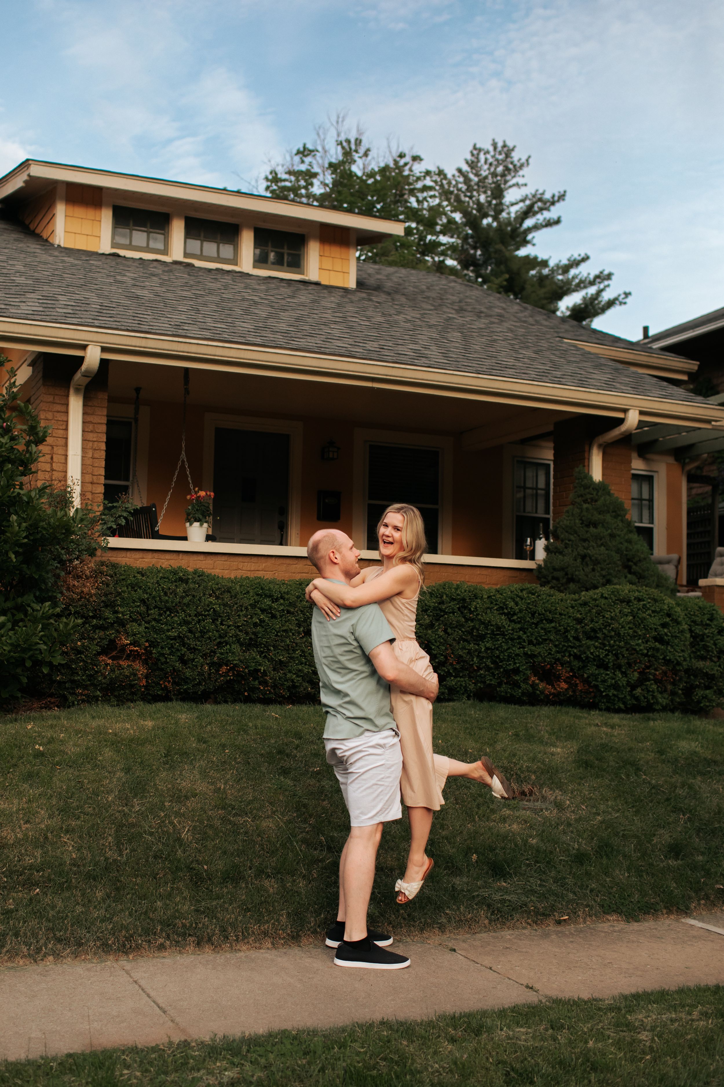 Two people embrace playfully on a lawn in front of a ranch-style house on a summer day.