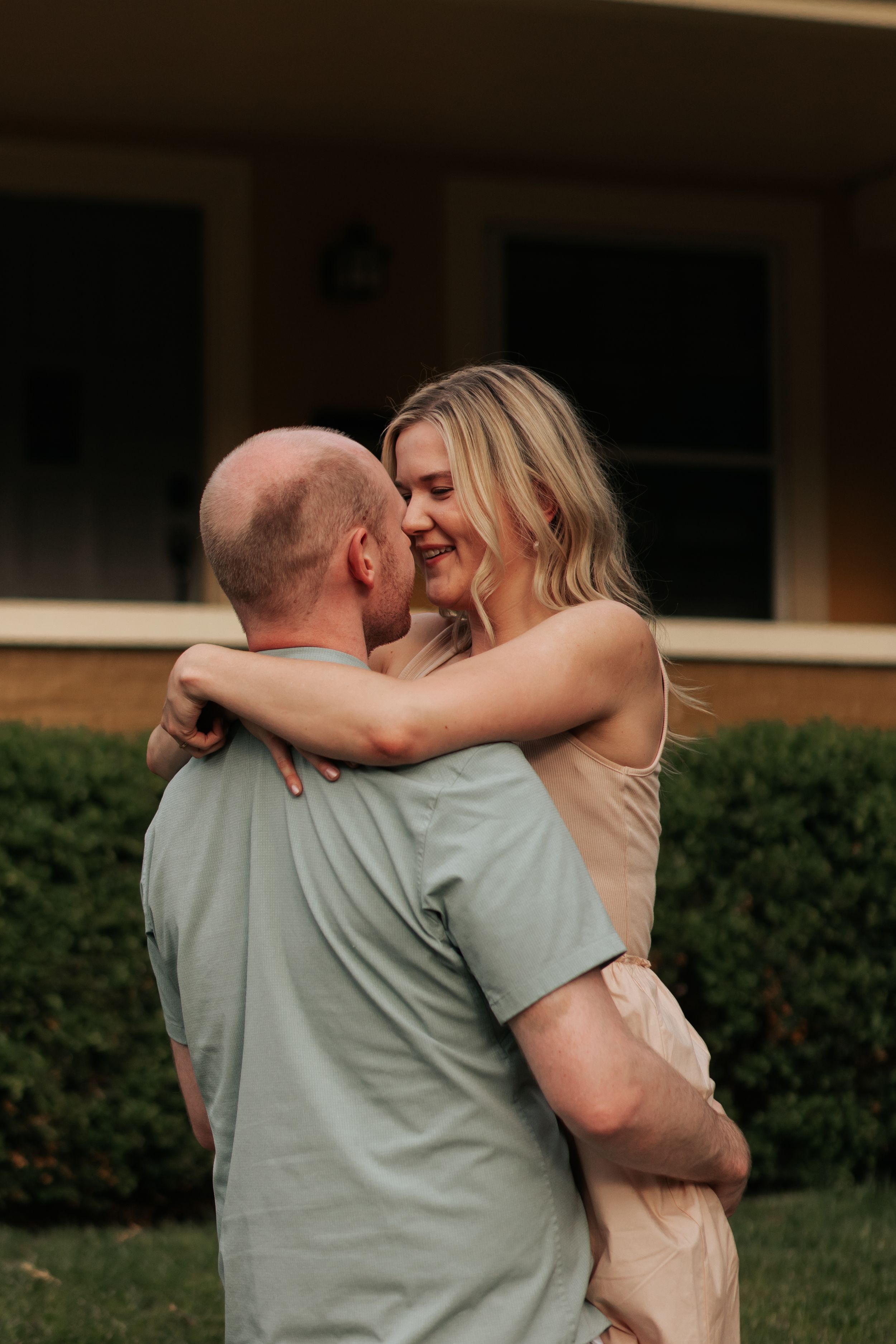 A loving couple embraces outdoors in front of a building with hedges, sharing an intimate moment together.