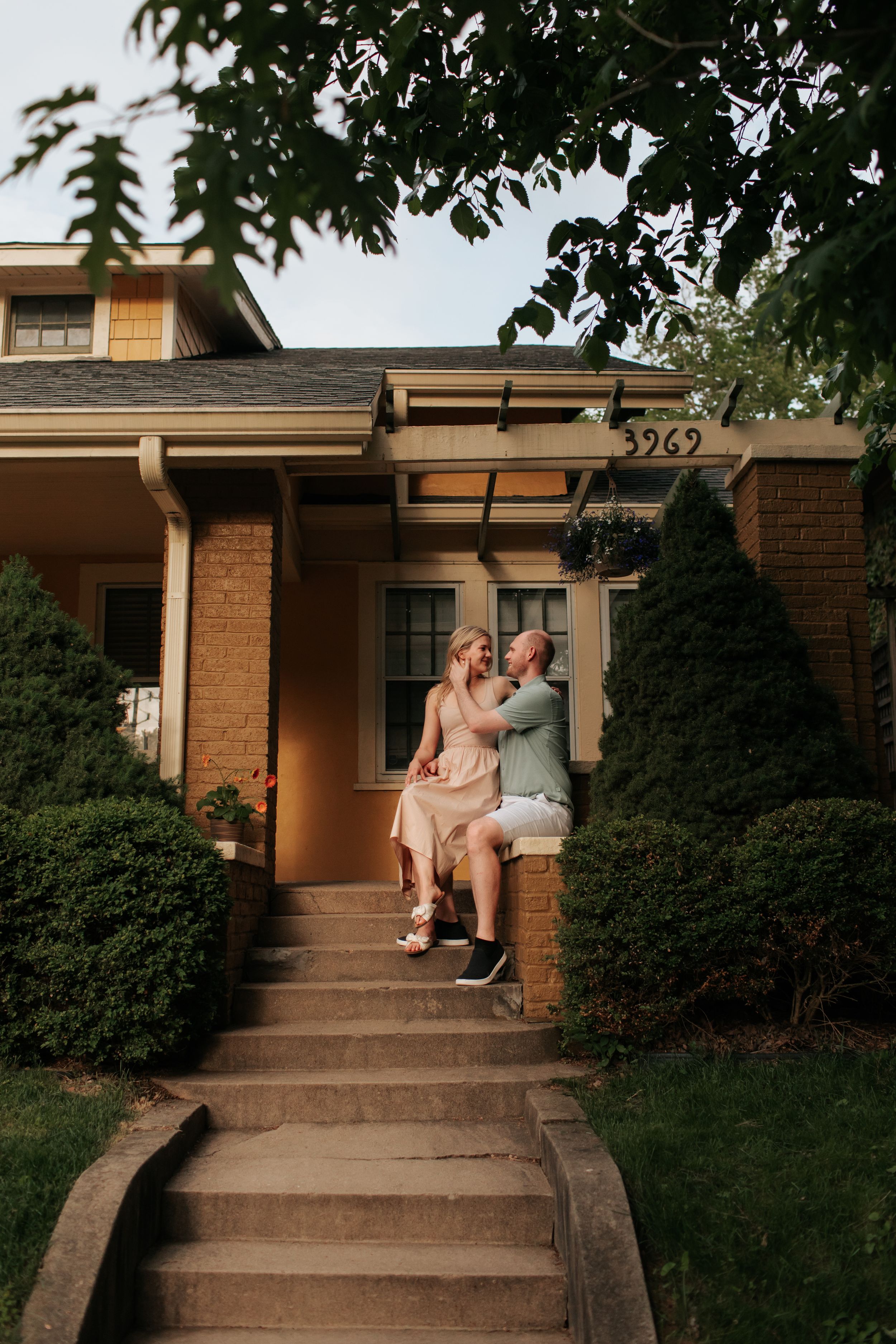 A couple sits together on the front steps of a brick house with shrubs and trees framing the entrance.