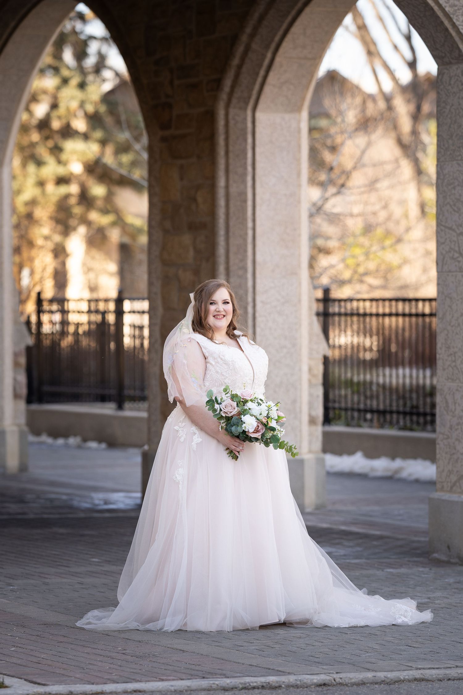 A bride in a flowing white wedding dress poses gracefully under a columned archway during a bridal photo session.