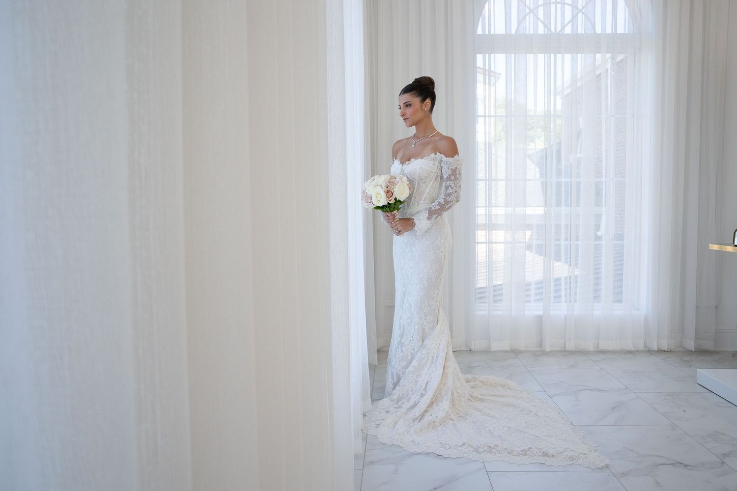 A bride in an elegant white gown stands by sheer curtains in a bright, minimalist room.