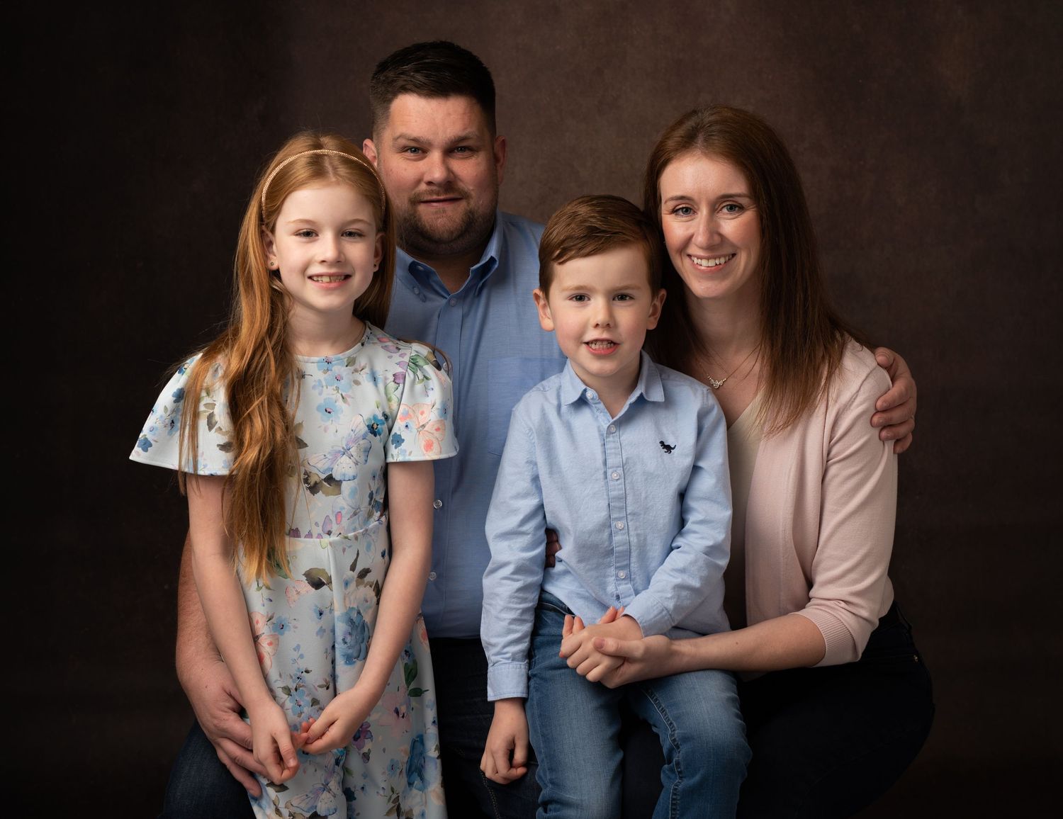 A family poses together in coordinated blue and neutral outfits against a dark studio backdrop.