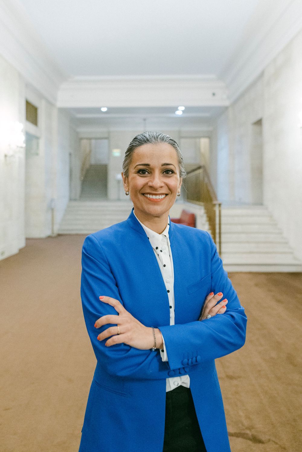 Professional in bright blue blazer and white shirt stands confidently in elegant hallway with marble details.