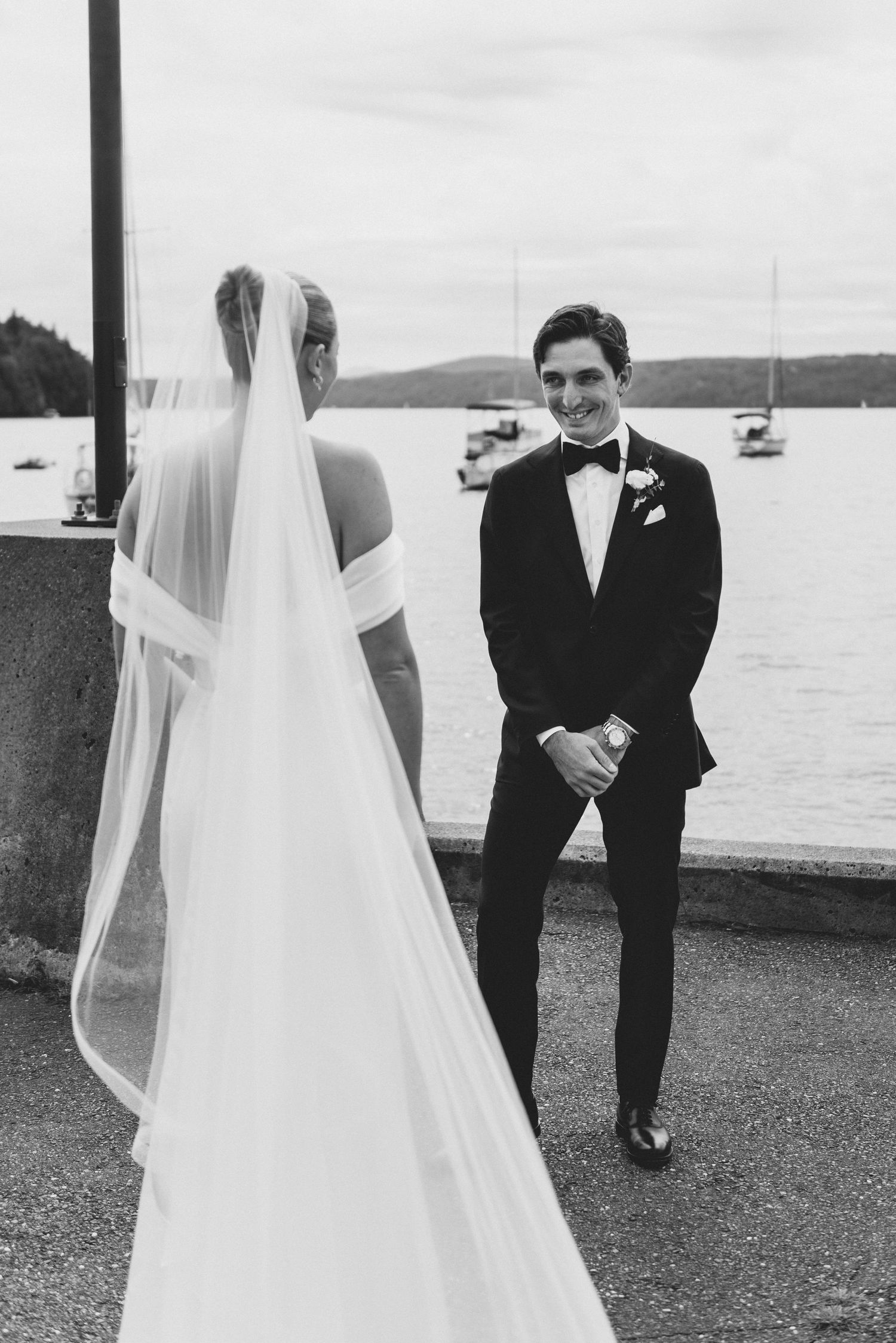 A bride in a flowing veil and groom in tuxedo share a candid moment by the waterfront with sailboats in the background.