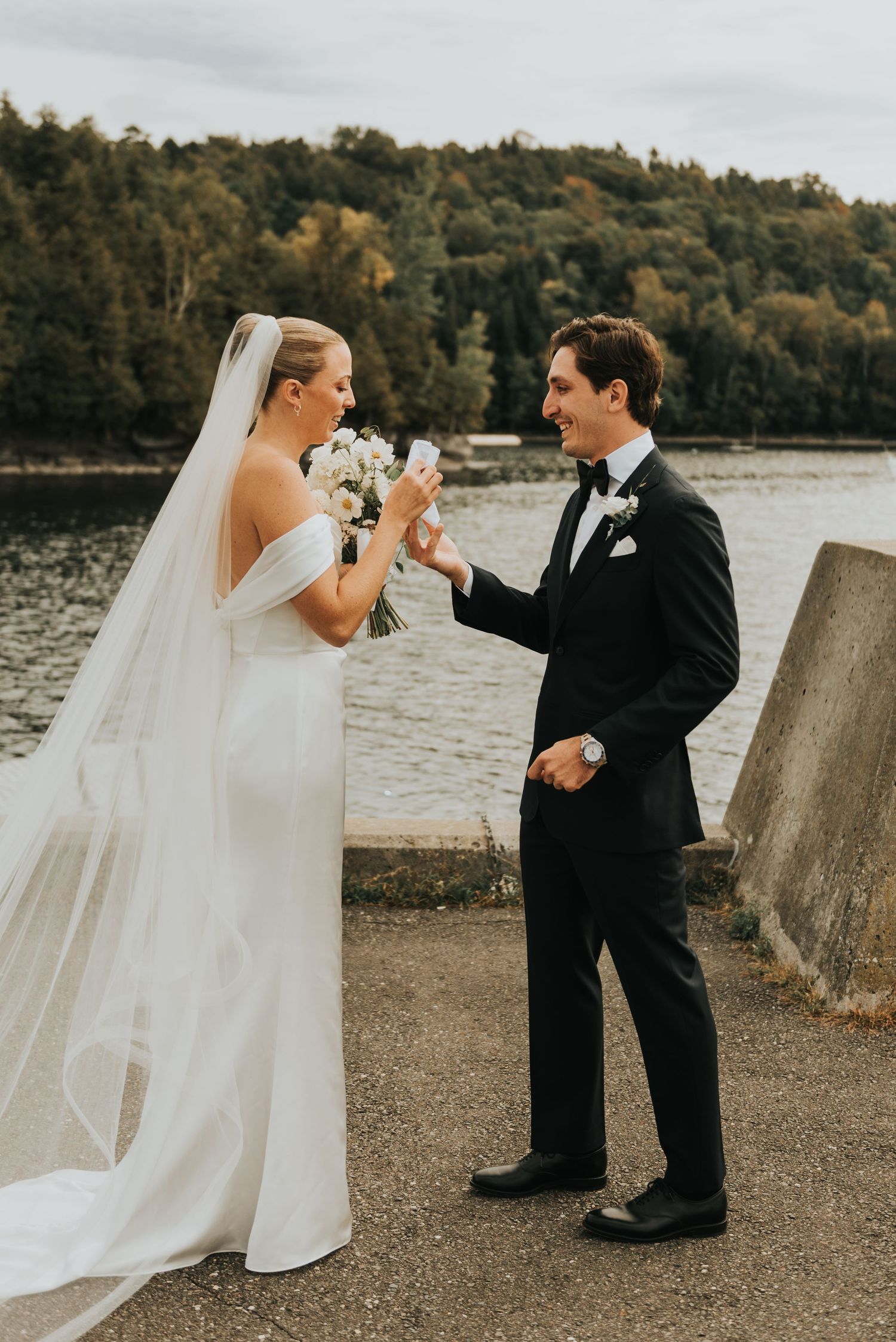 A bride and groom share a playful moment toasting by the water during their wedding photoshoot.