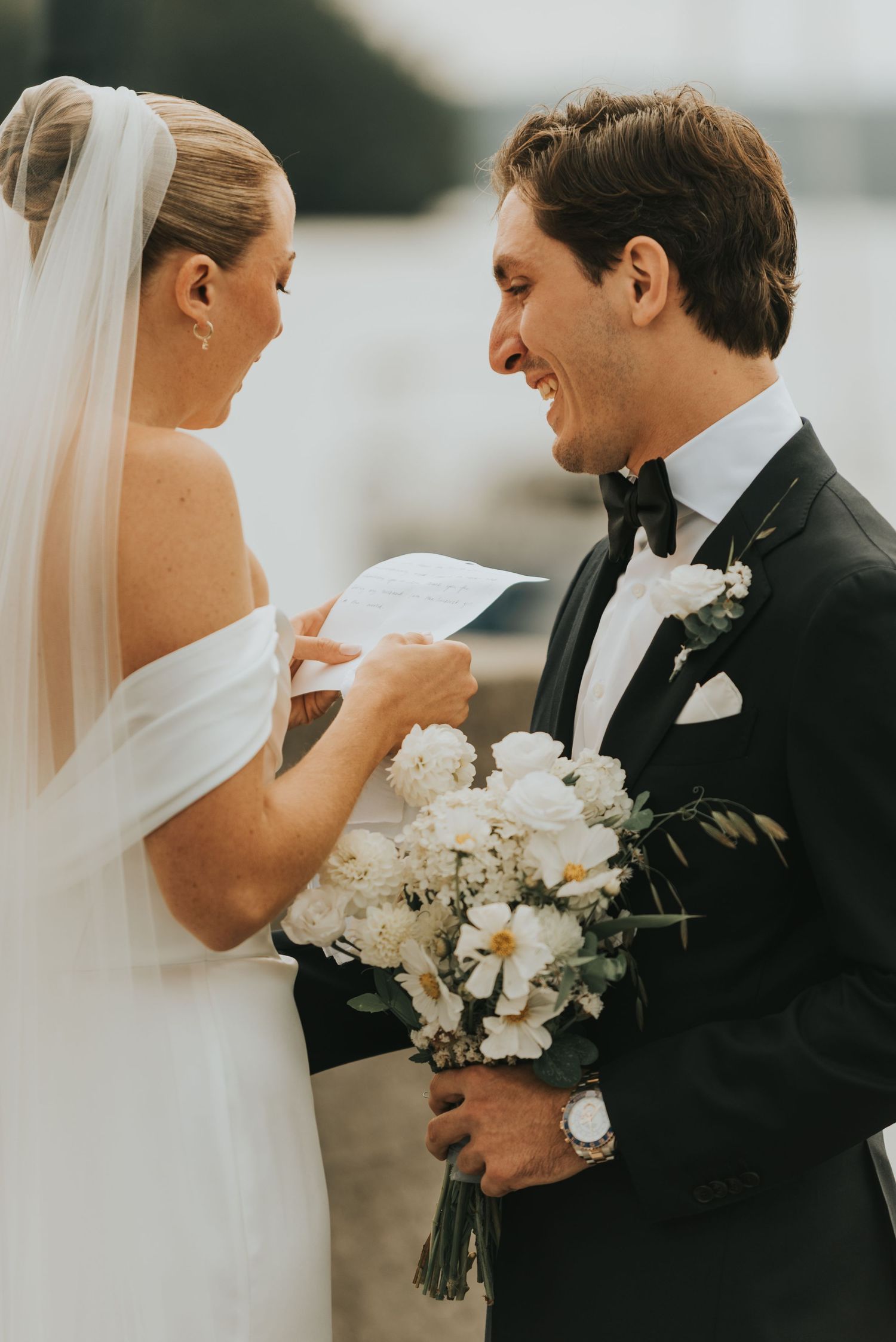A wedding couple shares an intimate moment as the bride reads vows while holding a white floral bouquet.