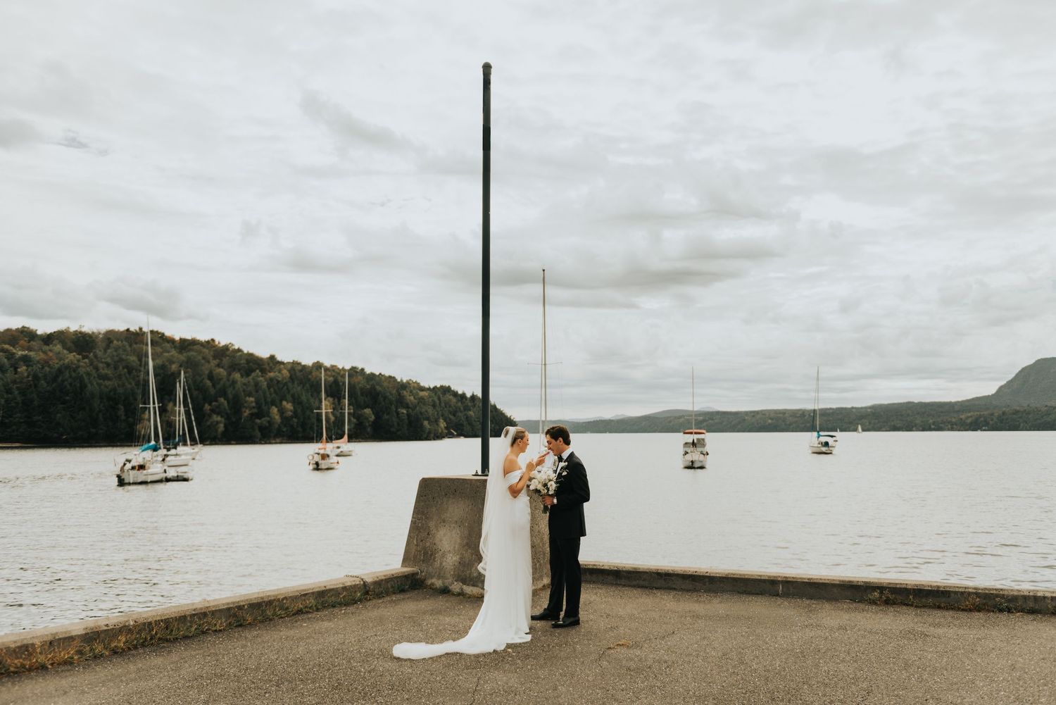Scenic waterfront wedding ceremony with sailboats anchored in the misty harbor under cloudy skies.