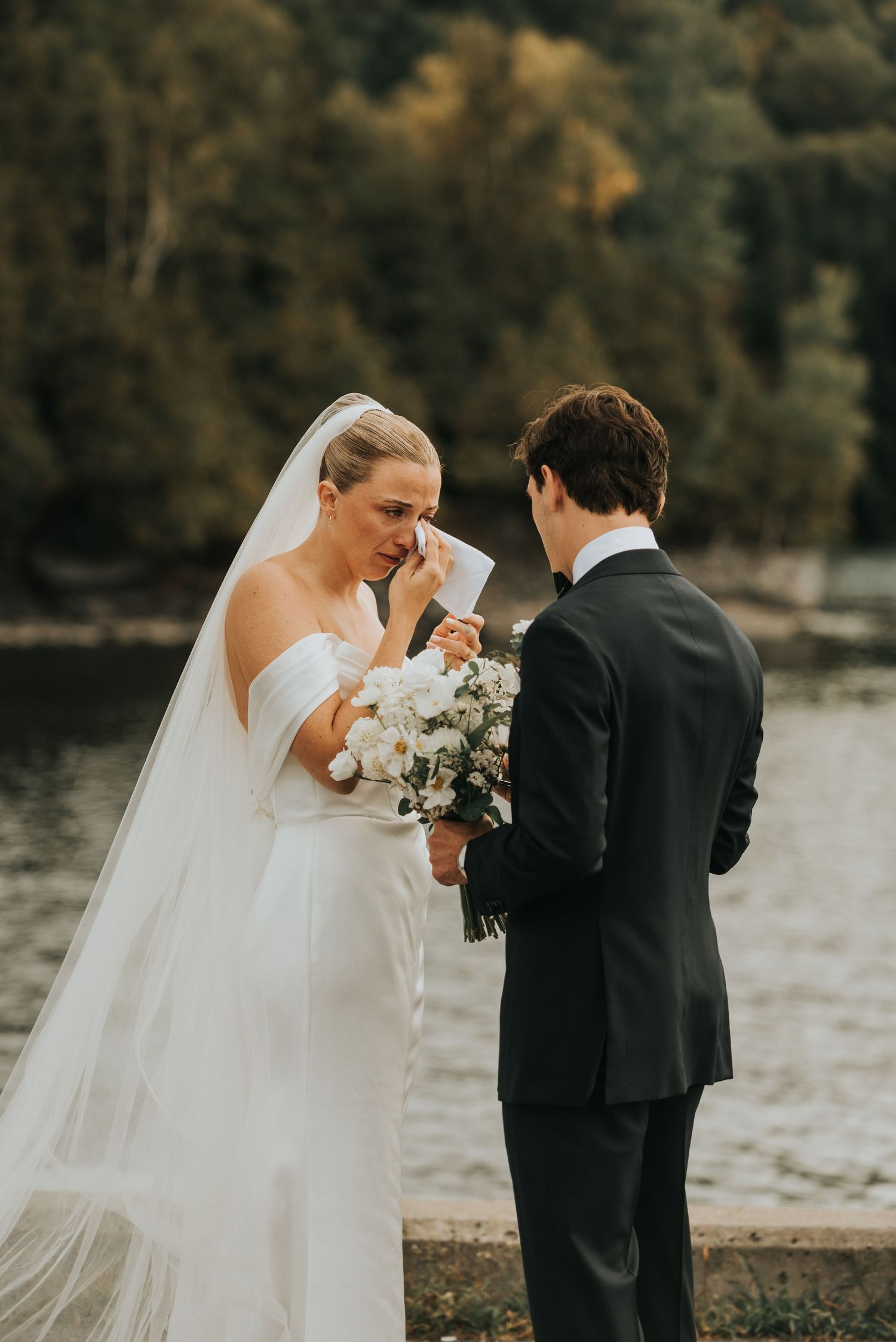 Wedding couple in black and white attire share an intimate moment with bouquet by the water.
