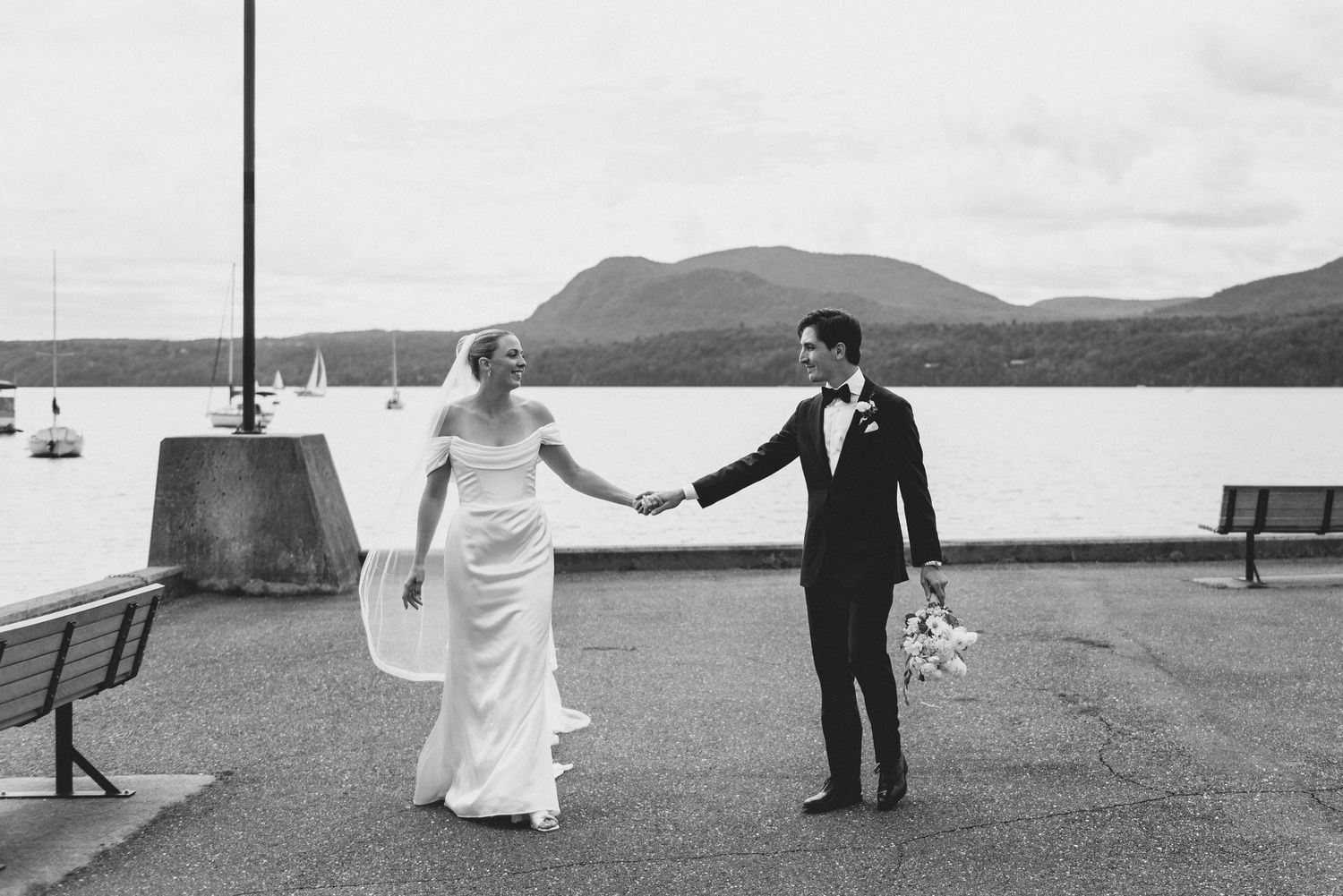 A couple in wedding attire hold hands while walking along a waterfront promenade with mountains in the background.