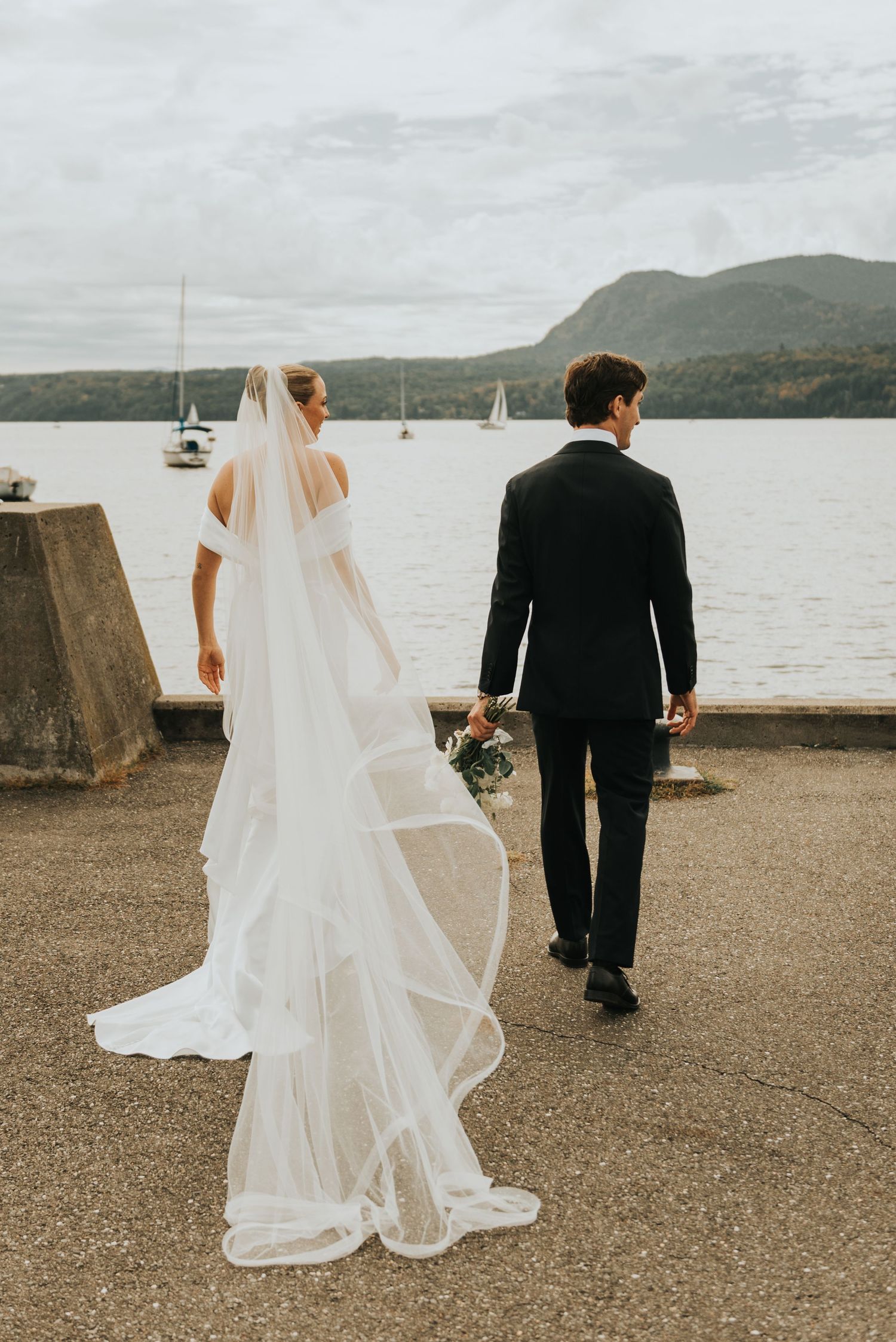 A bride and groom walk along a waterfront path with mountains and sailboats in the distance.