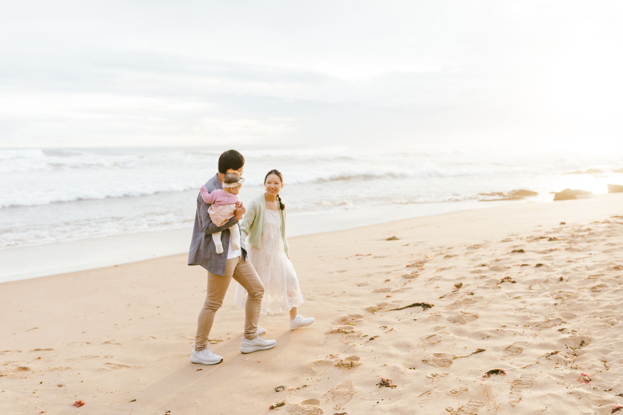 Couple walking along a sandy beach at sunset holding hands with ocean waves in the background.