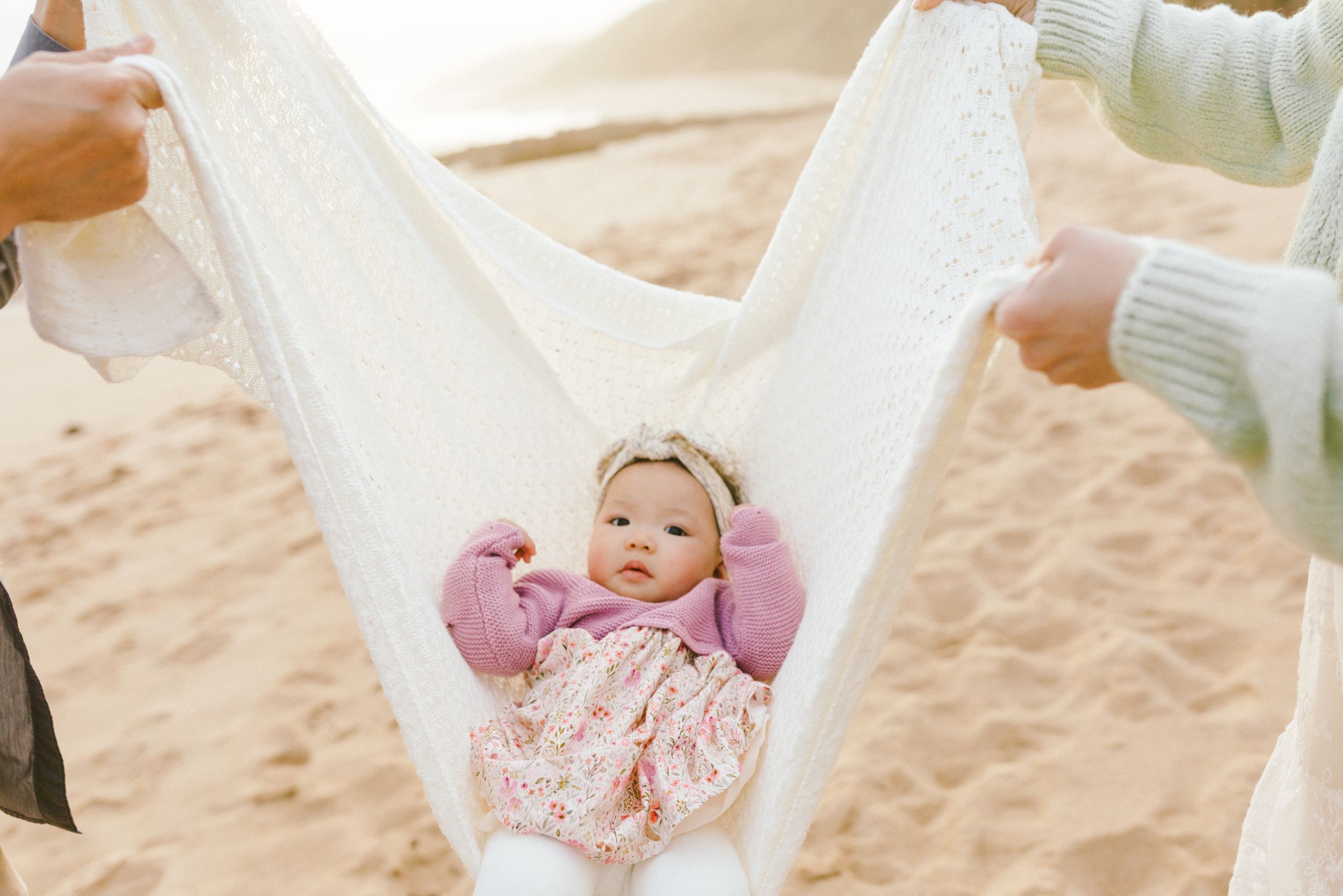A white hammock swings gently on a sandy beach with palm trees and ocean waves visible in the background.