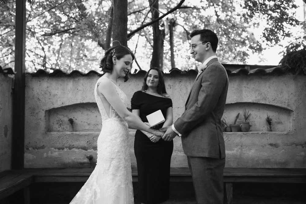 Black and white photo of a wedding ceremony taking place under a rustic wooden pergola with stone walls.