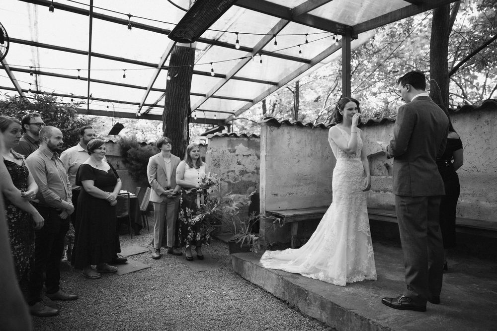 Intimate wedding ceremony taking place under a wooden pergola with stone wall backdrop.