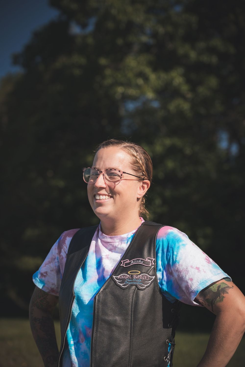 Person in tie-dye shirt and leather vest smiles outdoors on a sunny day.