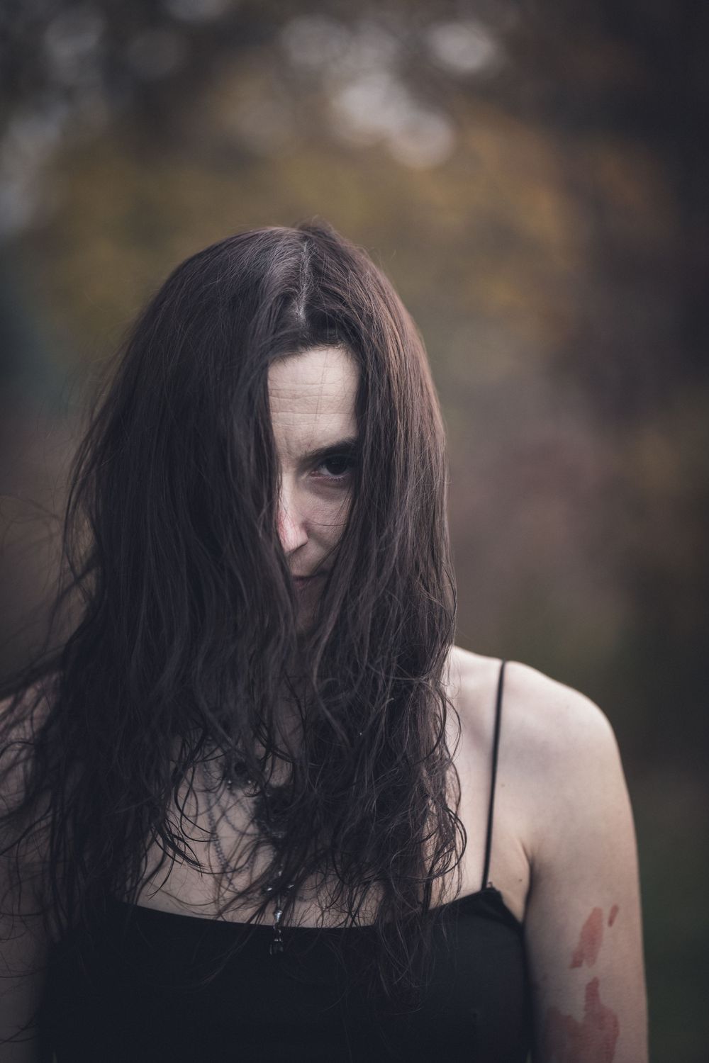 Artistic dark portrait with long black hair against blurred natural background.