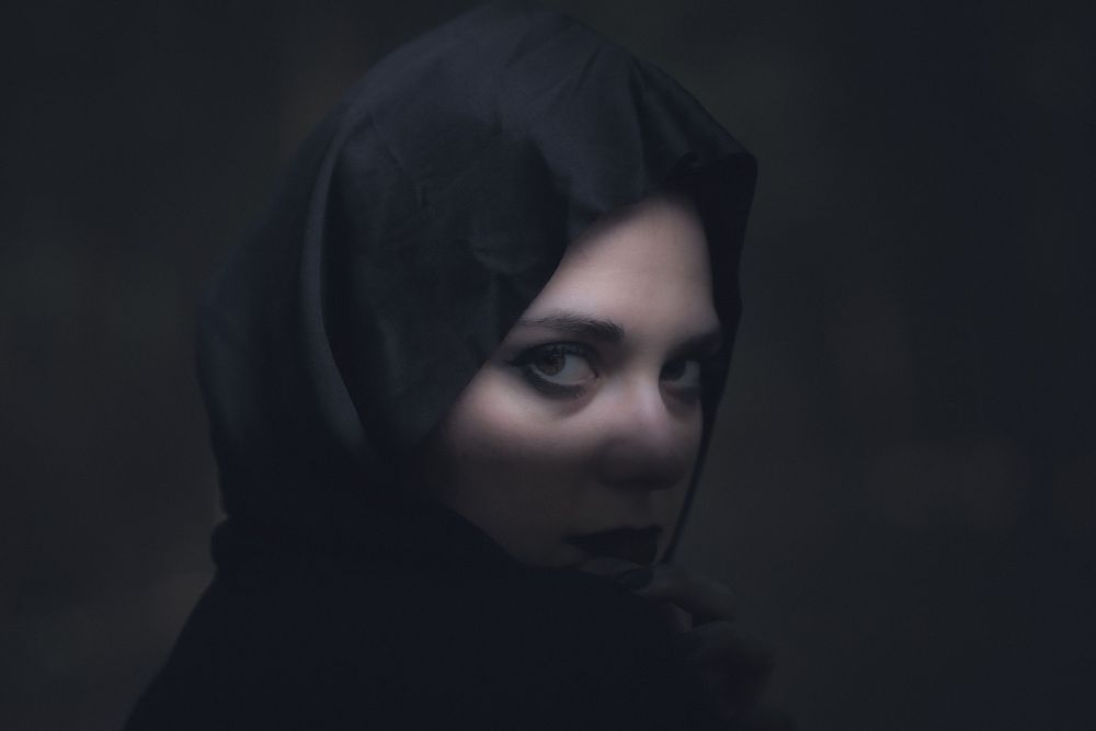 Atmospheric portrait of a figure in a black hijab against a dark moody background.