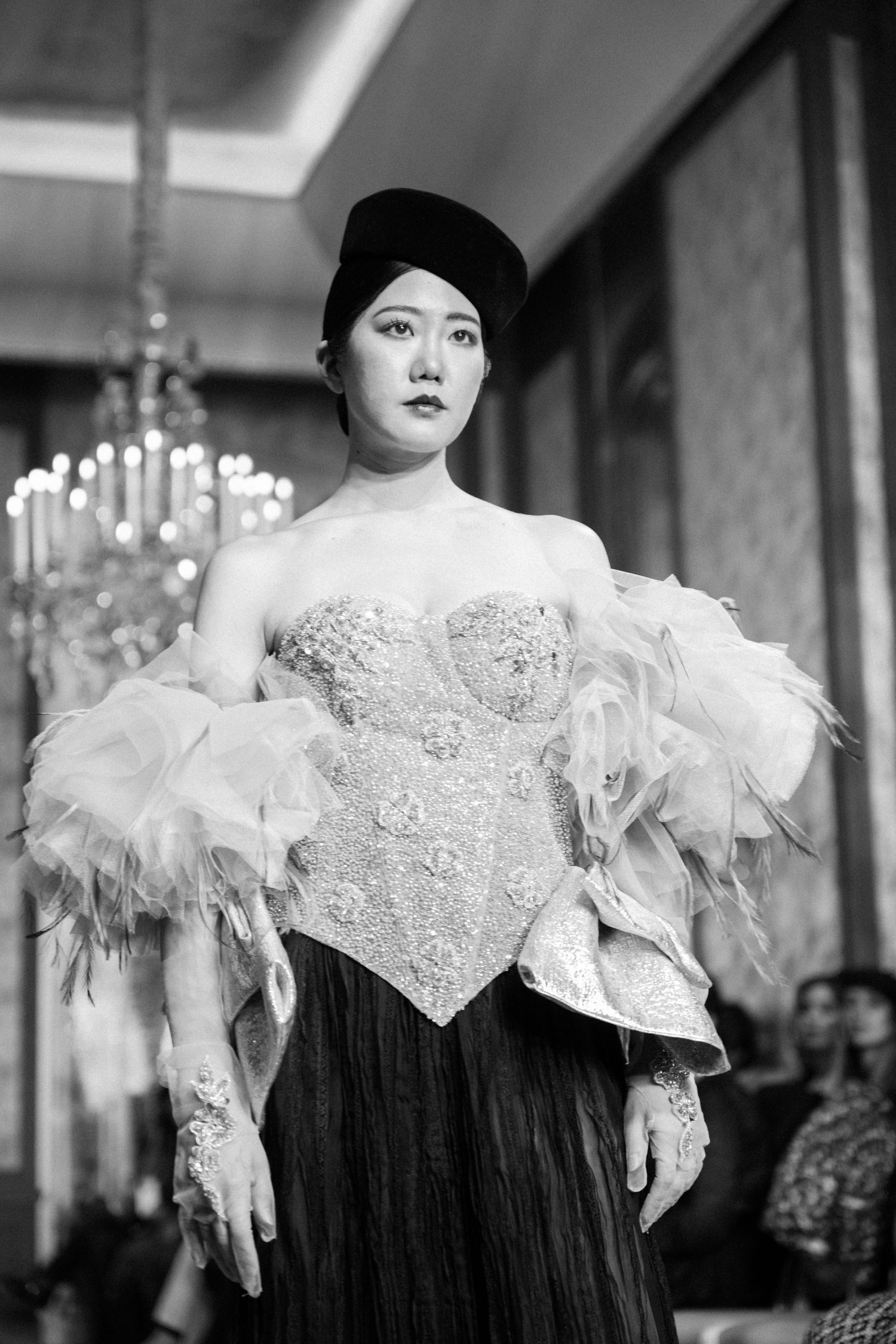 A fashion model wearing an ornate black and white gown with feathers poses in front of a chandelier.