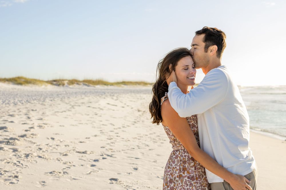A romantic couple embraces on a sunny beach with white sand and ocean waves in the background.