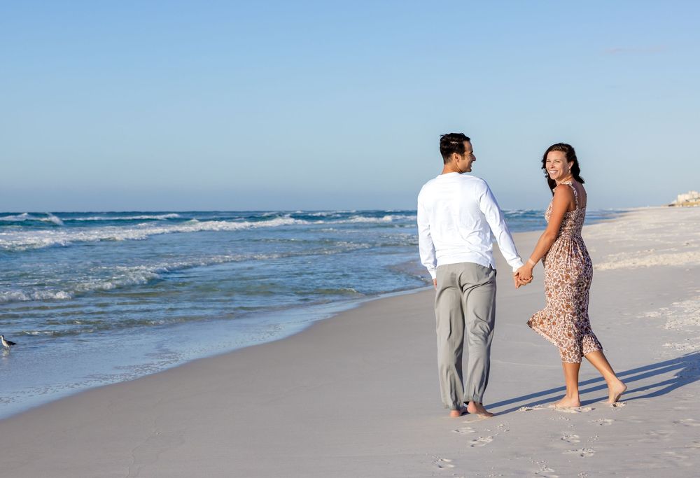 A couple walks hand in hand along a serene beach shoreline with gentle waves rolling in on a sunny day.