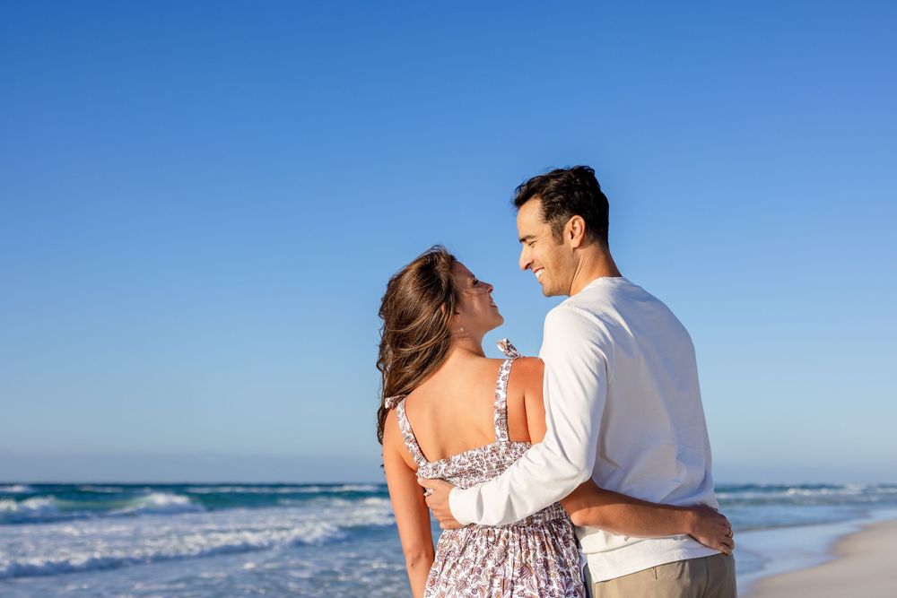 Romantic couple embracing while looking at ocean waves on sunny beach with blue sky.