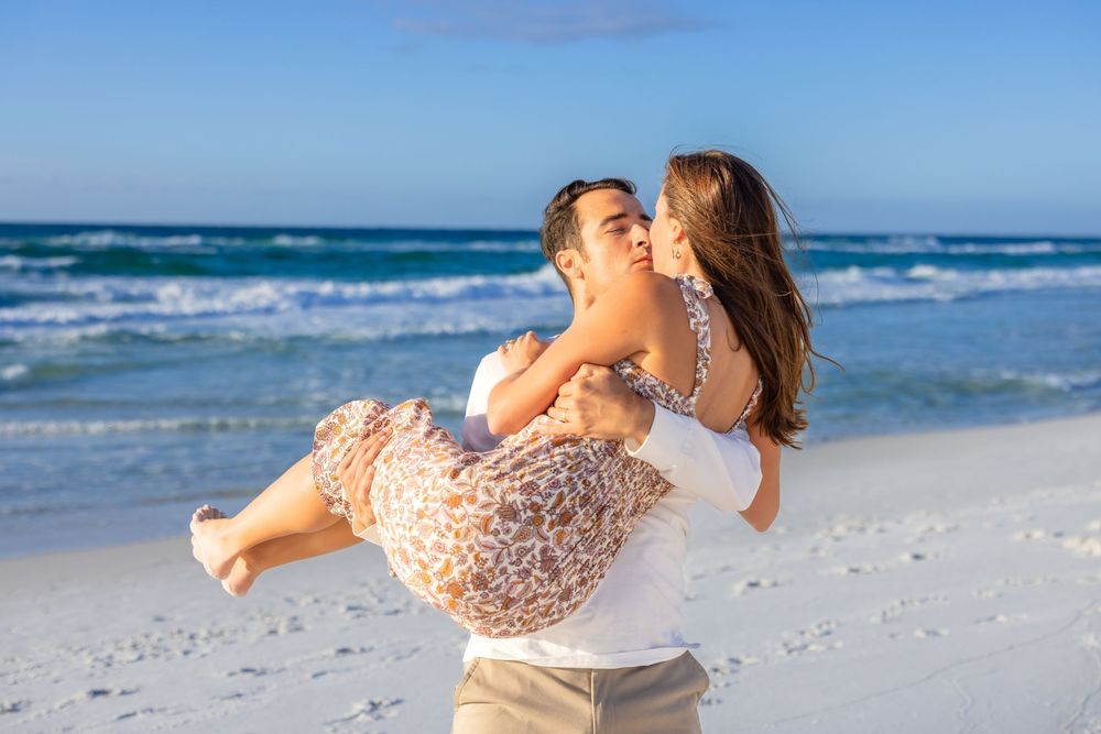A couple embraces romantically on a white sandy beach with turquoise ocean waves in the background.