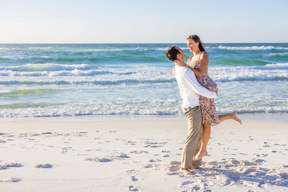 Couple in casual attire embracing on a sunny beach with waves crashing in the background.