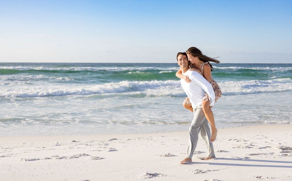 Woman in white dress walking along a pristine beach with turquoise waters and white sand on a sunny day.