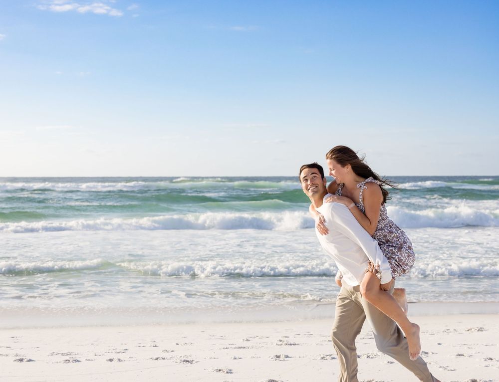 Two people share a joyful moment together while walking on a sunny white sand beach with crashing waves.