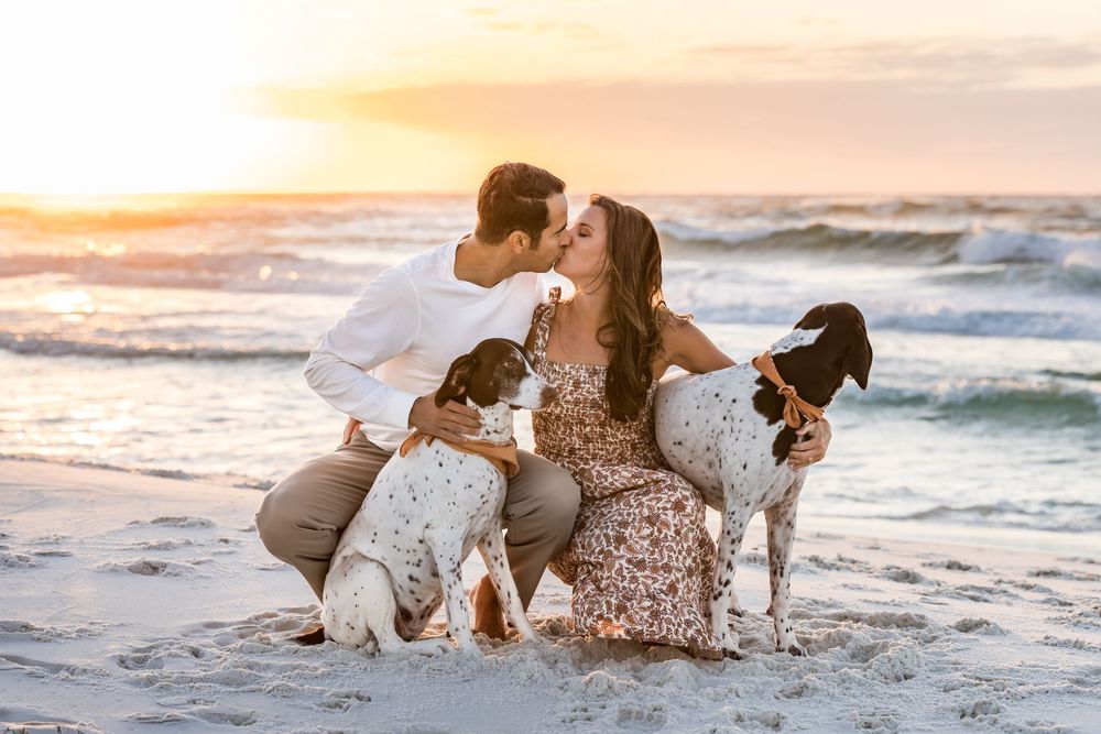 A couple and their dog share tender moments on a sandy beach at sunset with ocean waves in the background.