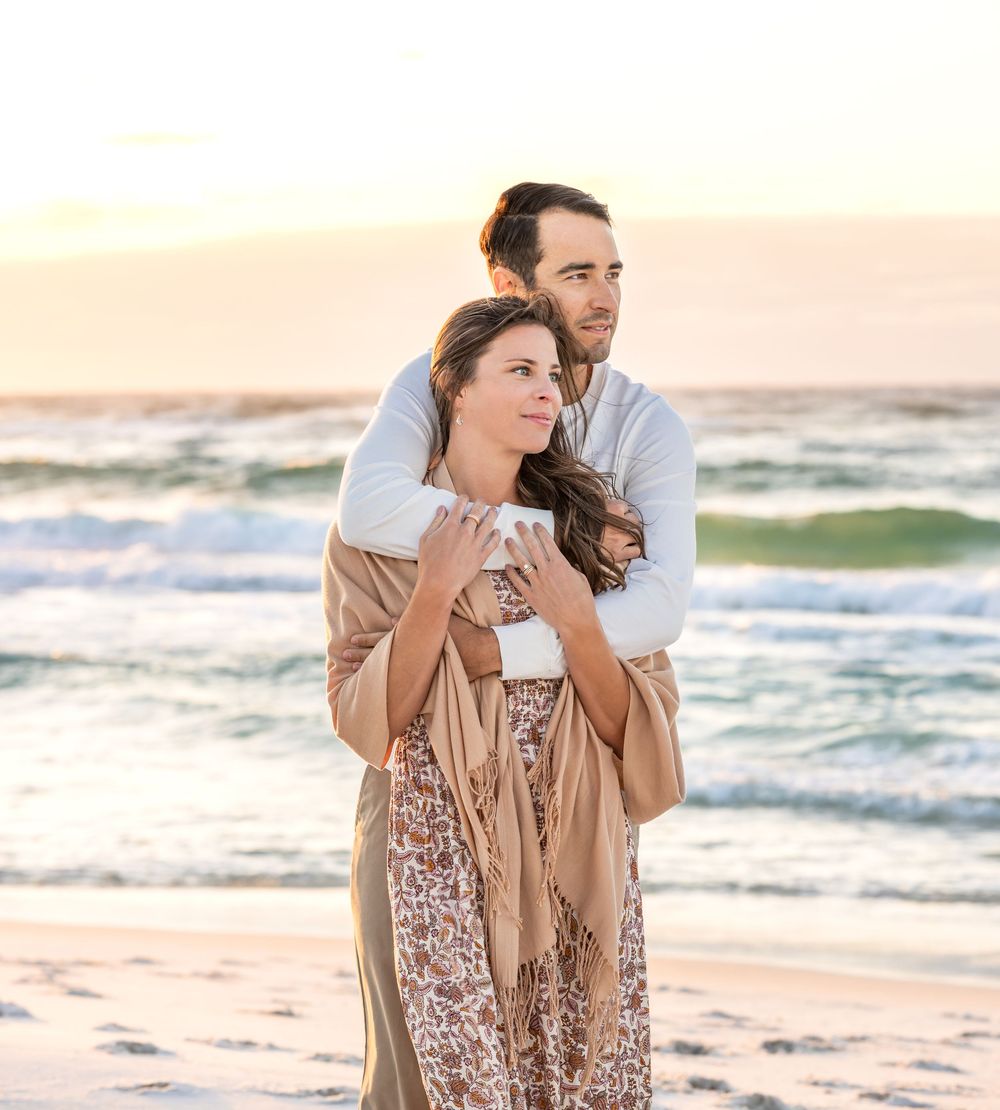 A couple embraces on a beach at sunset with ocean waves rolling in the background.