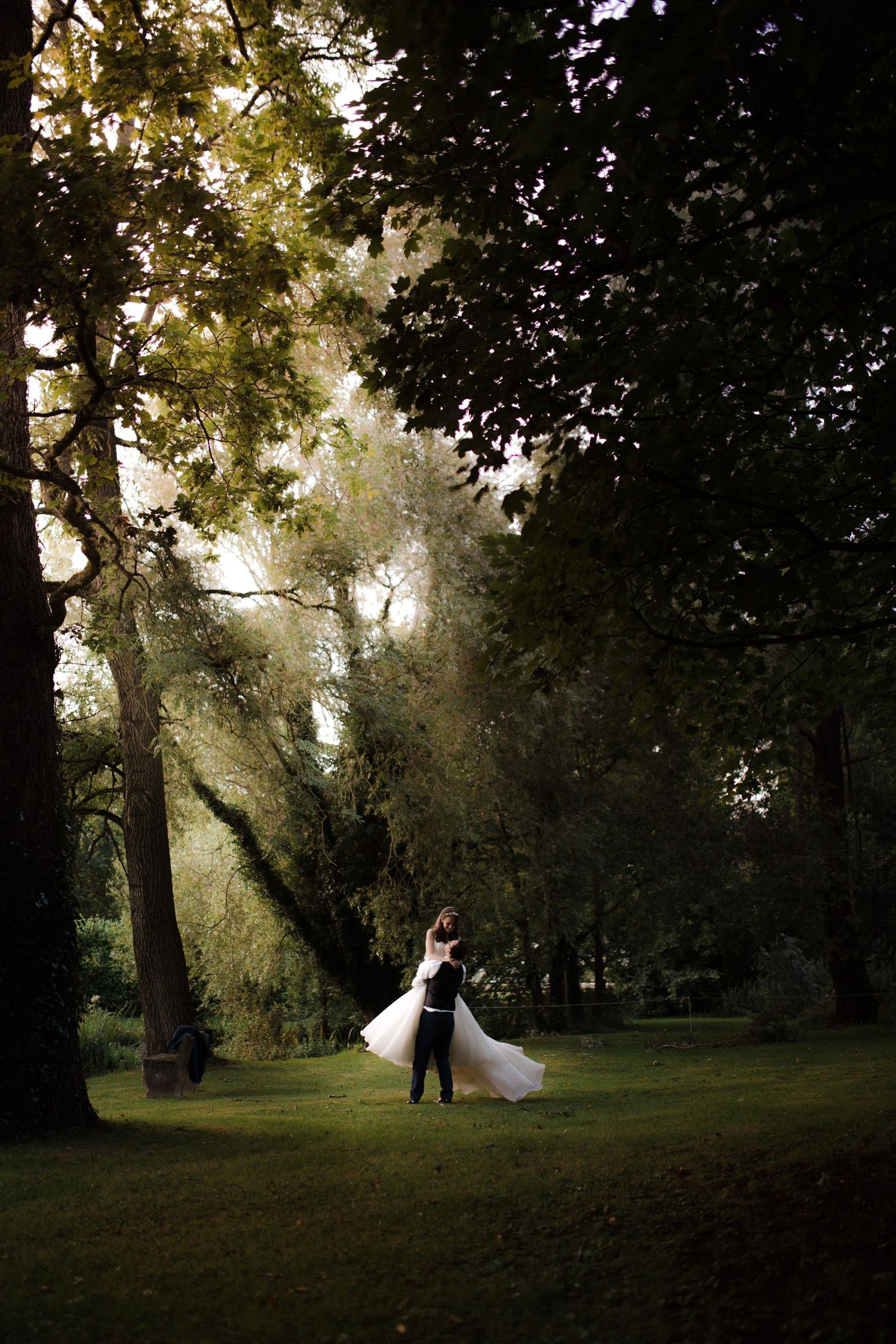 A couple in wedding attire walks through a misty forest with sunlight streaming through the trees.