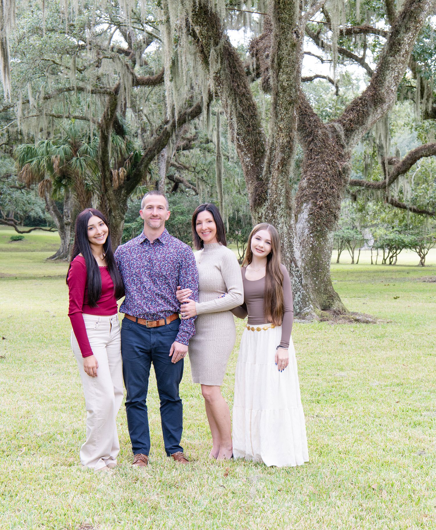Family of four posing together outdoors under large moss-covered oak trees on a green lawn.