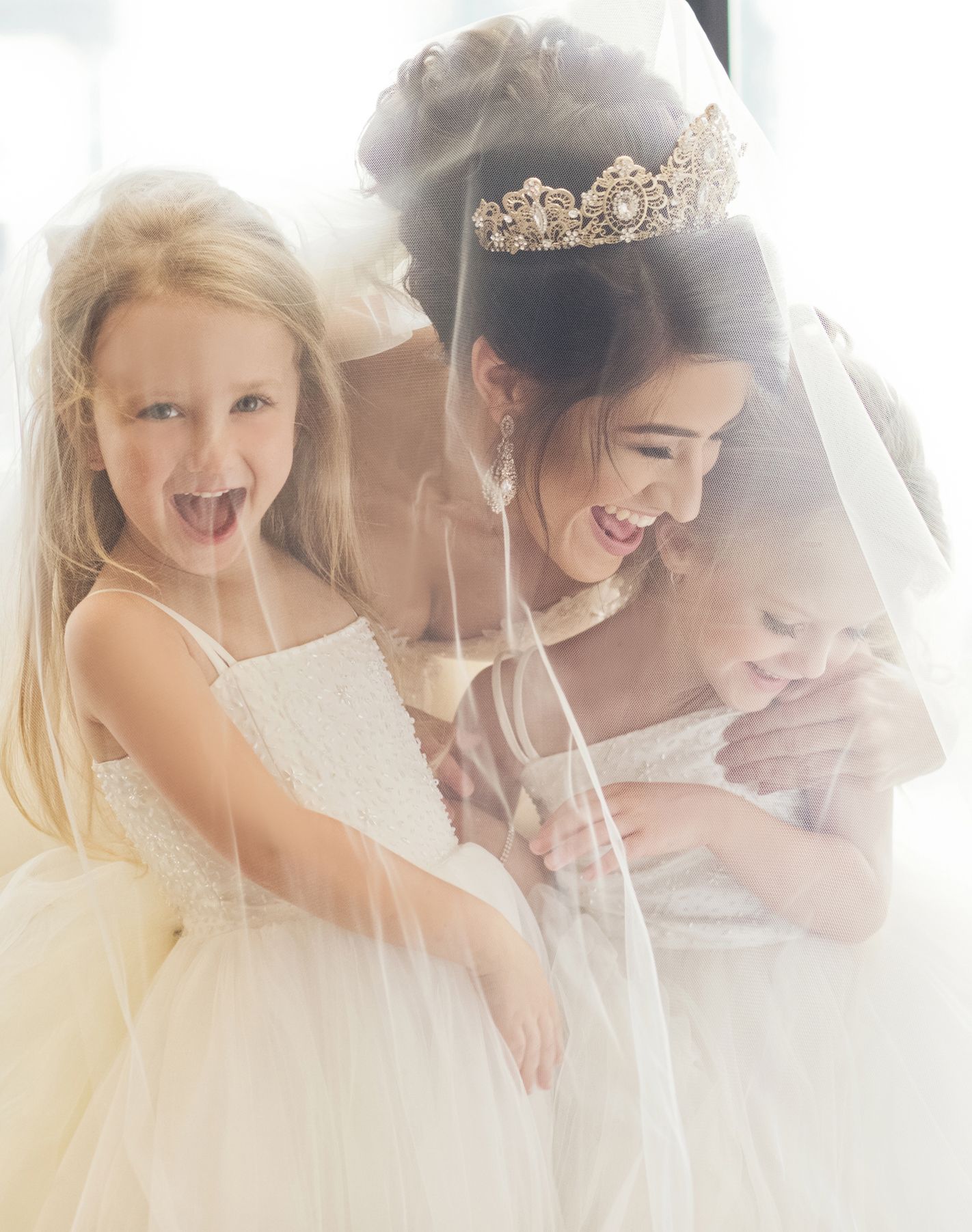 Smiling bride wearing tiara and veil shares joyful moment with two young flower girls in white dresses.