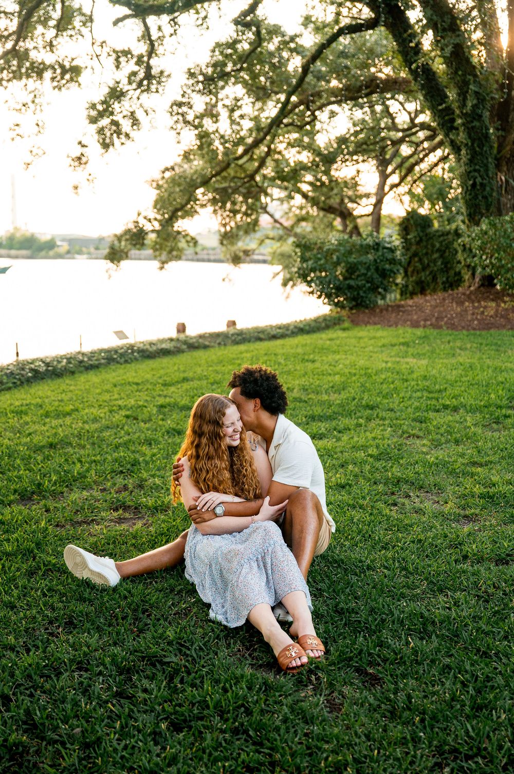 A couple relaxes on a grassy hillside beneath trees during sunset.