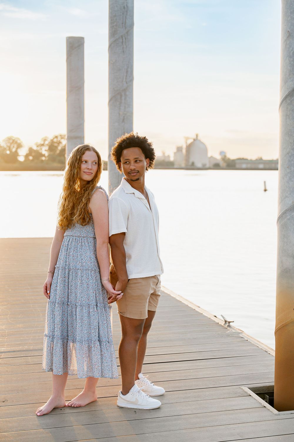 A couple in coordinated outfits stands together on a wooden pier overlooking the water at sunset.