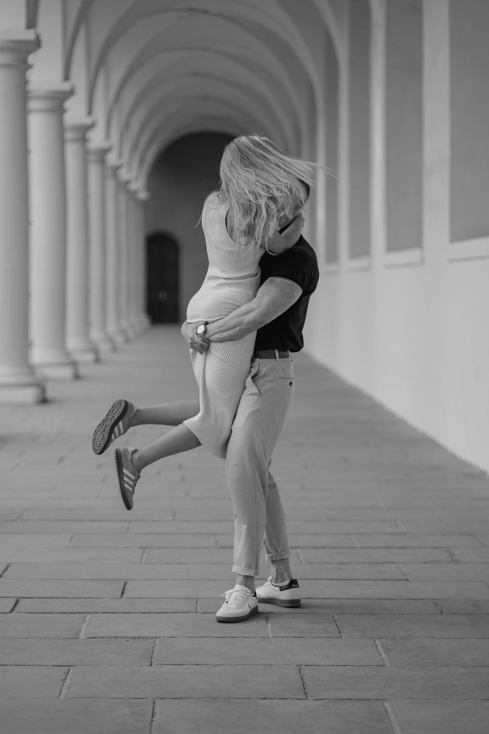 A romantic black and white photo of a couple embracing in an arched colonnade hallway.