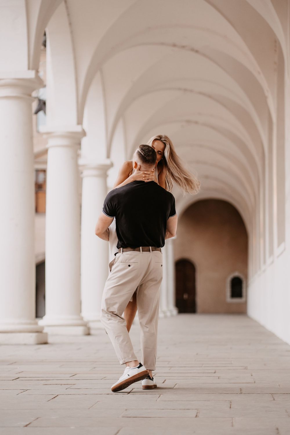 A couple dances romantically in an elegant white archway corridor with classical architecture and soft lighting.